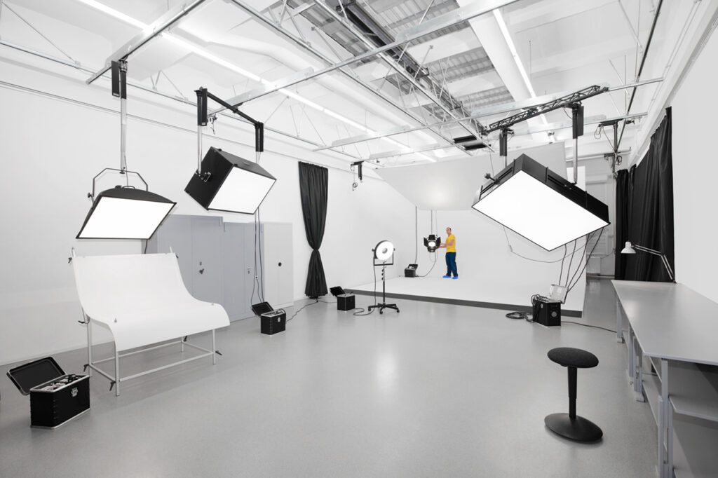 Ein geräumiges, modernes Fotostudio mit großen Softbox-Leuchten, einem weißen Hintergrund und einer Person in farbenfroher Kleidung, die ganz hinten steht. Der Raum ist aufgeräumt, übersichtlich und gut beleuchtet.